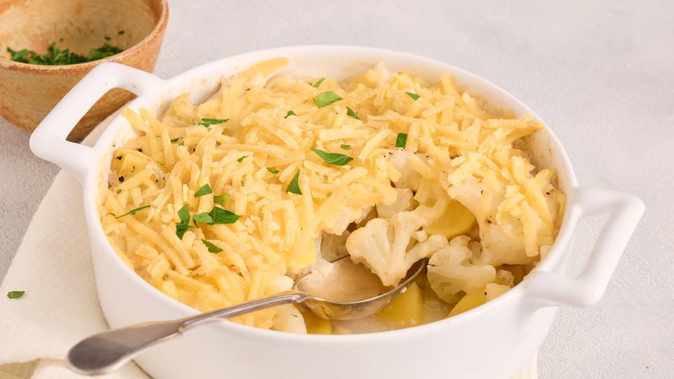 Ein ofenfrisches Blumenkohl-Kartoffel-Gratin in einer weißen Auflaufform: zarte Blumenkohlröschen und Kartoffelscheiben, überbacken mit goldbraunem geriebenem Käse und frisch gehackter Petersilie. Serviert mit einem Löffel zum Portionieren.