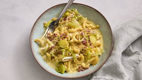 Ein Teller mit cremigen Spätzle, verfeinert mit fein geschnittenen Lauchringen und knusprigen Speckwürfeln. Das Gericht wirkt herzhaft und deftig.