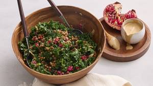 Ein frischer Grünkohlsalat mit Linsen und Granatapfelkernen in einer hölzernen Schüssel, dazu eine Schale mit cremigem Dressing und einer halbierten Granatapfelfrucht.