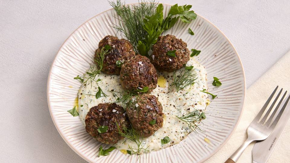Auf einem weißen Porzellanteller mit einem Reliefmuster wird ein Gericht mit Fleischbällchen auf einer weißen Joghurt-Soße, garniert mit frischen Kräutern serviert. Neben dem Teller liegt eine cremefarbene Stoffserviette mit Gabel und Messer.