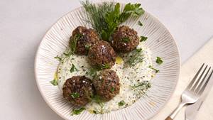 Auf einem weißen Porzellanteller mit einem Reliefmuster wird ein Gericht mit Fleischbällchen auf einer weißen Joghurt-Soße, garniert mit frischen Kräutern serviert. Neben dem Teller liegt eine cremefarbene Stoffserviette mit Gabel und Messer.