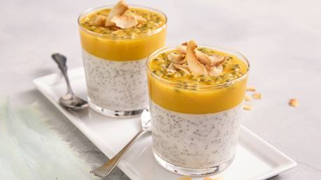 Auf einem hellen Untergrund ist ein rechteckiger weißer Teller mit zwei Gläsern mit einem geschichteten Dessert. Die untere Schicht besteht aus Chia-Samen-Pudding, die obere aus einer gelben Mango-Maracuja-Fruchtsoße, garniert mit gerösteten Kokoschips. Neben jedem Glas liegt ein Teelöffel mit langem Stil.