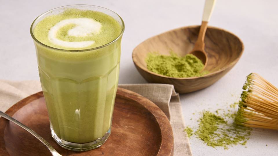 Auf einem hellen Untergrund liegt eine hellbraune Stoffserviette auf dem ein runder dunkler Holzteller steht. Auf dem Holzteller steht ein hohes Glas mit einem grünen Getränk, einem Matcha-Latte. Auf dem Teller liegt ebenfalls ein Teelöffel mit grünem Matchapulver. Im Hintergrund ist eine kleine Holzschüssel mit mehr Matchapulver.