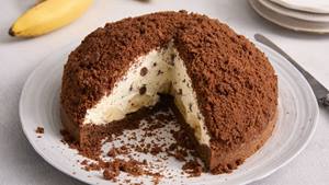 Auf einem hellen Untergrund ist ein großer weißer Porzellanteller mit einem Kuchen. Der Kuchen ist angeschnitten und etwa ein Drittel wurde entnommen, das Innere des Kuchens ist zu sehen. Der Kuchen, der sogenannte Maulwurfkuchen, hat einen Boden aus Schokoteig, eine Sahne-Schokotropfen-Banane-Füllung und ist nochmal mit Schokoteig bestreut. Im Hintergrund ist eine Banane und weitere Teller.  