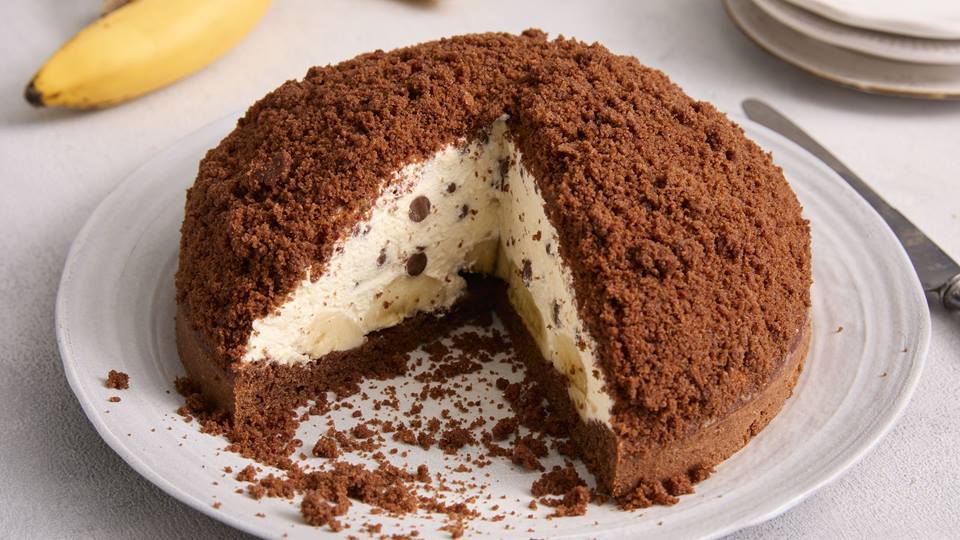 Auf einem hellen Untergrund ist ein großer weißer Porzellanteller mit einem Kuchen. Der Kuchen ist angeschnitten und etwa ein Drittel wurde entnommen, das Innere des Kuchens ist zu sehen. Der Kuchen, der sogenannte Maulwurfkuchen, hat einen Boden aus Schokoteig, eine Sahne-Schokotropfen-Banane-Füllung und ist nochmal mit Schokoteig bestreut. Im Hintergrund ist eine Banane und weitere Teller.  