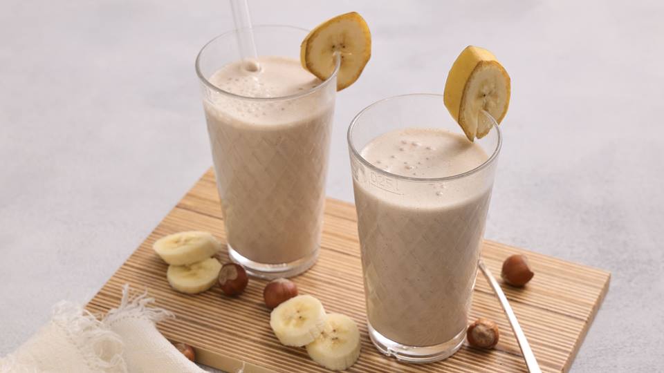 Auf einem hellen Untergrund liegt ein helles Holzbrett. Auf diesem stehen zwei höhere Gläser mit einem cremefarbenen Bananen-Milchshake. Die Gläser sind mit einem Stück Banane mit Schale garniert. Auf dem Holzbrett liegen ebenso vier Stück Bananen ohne Schale und Haselnüsse mit Schale.