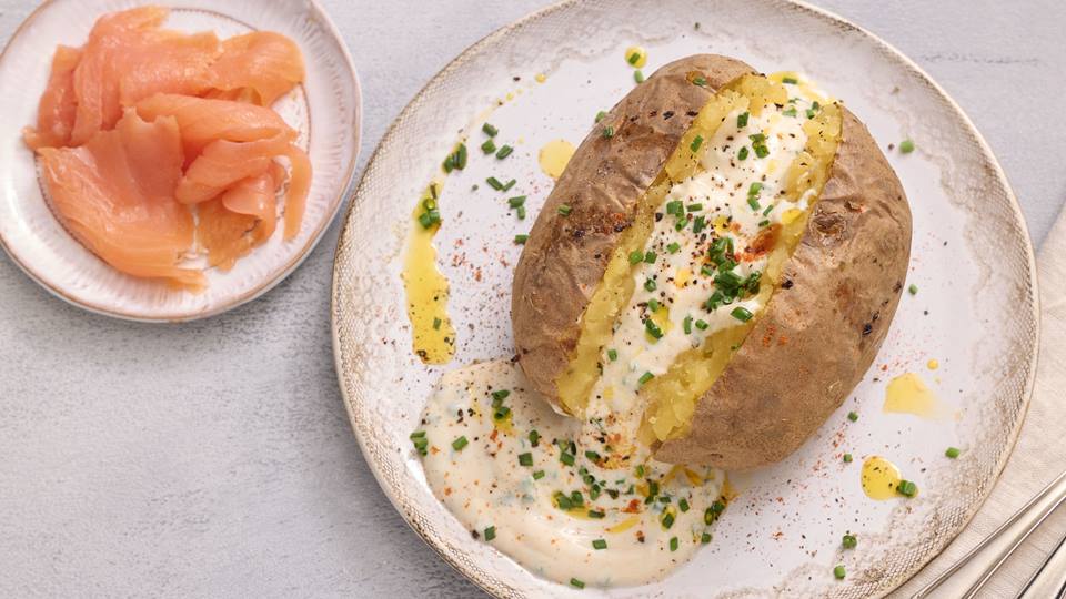 Auf einem hellen Untergrund ist ein hellbeiger Teller mit einer aufgeschnittenen Ofenkartoffeln. Im Schnitt der Kartoffel befindet sich eine weiße Soße, die mit Kräutern garniert ist. Links steht ein weiterer kleiner weißer Teller mit Räucherlachs.