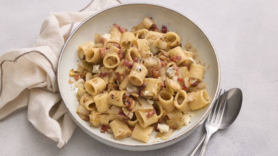Auf einem hellen Untergrund ist ein tieferer hellbeiger Teller mit Nudeln in Form von Röhrchen. Zwischen den Nudeln sind Speckwürfel und geschnittener Chicoree. Rechts neben dem Teller liegen eine Gabel und ein Esslöffel sowie links eine helle Stoffserviette.