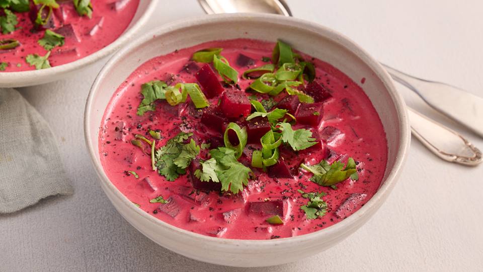 Auf einem hellen Untergrund ist in einer weißen Schüssel eine rotfarbene Suppe aus Rote-Bete, garniert mit krauser Petersilie. Neben der Schüssel liegt ein Esslöffel und ein weitere Schüssel mit Suppe ist im Anschnitt erkennbar.