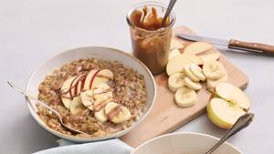 In einer weißen Porzellanschüssel ist ein Porridge garniert mit Apfelscheiben. Seitlich in der Schüssel steckt ein Teelöffel. Im Hintergrund liegt eine hellblaue Stoffserviette und ein Holz-Schneidebrett, beides ist nur im Anschnitt erkennbar.