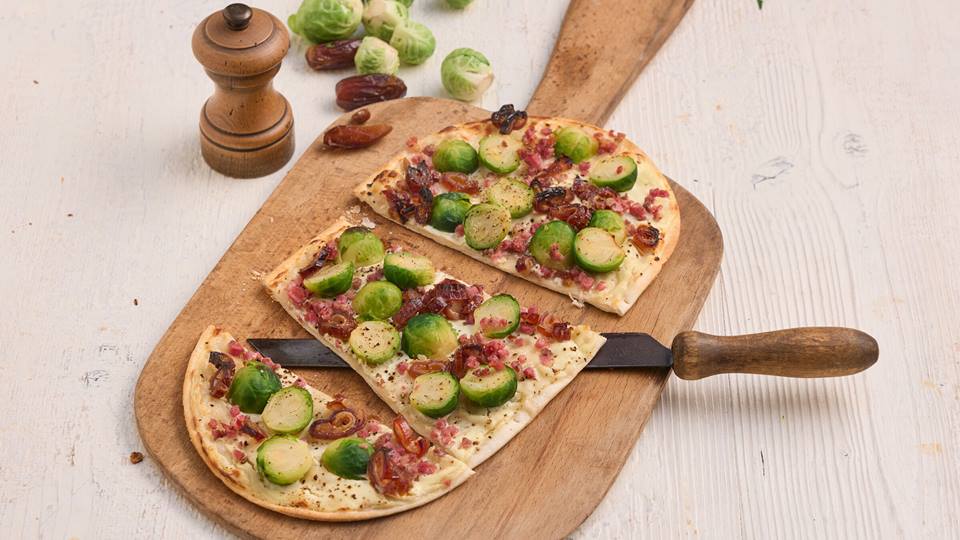 Ein Holzbrett mit einem Flammkuchen, belegt mit cremiger Basis, geschnittenen Datteln, halben Rosenkohlröschen und Speckwürfeln. Die Flammkuchen sind in Stücke geschnitten. Im Hintergrund sind frische Rosenkohlröschen, eine Pfeffermühle und Datteln zu sehen.