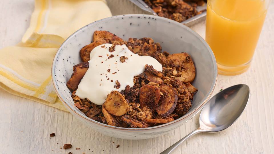 In einer Schüssel sind geröstete Bananen- und Kokosscheiben, Haferflocken und Mandeln, garniert mit einem Klecks Naturjoghurt. Neben der Schüssel steht ein Glas Orangensaft und liegt ein Löffel. Im Hintergrund ist ein Backbleck mit dem restlichen Kokos-Bananen-Granola.