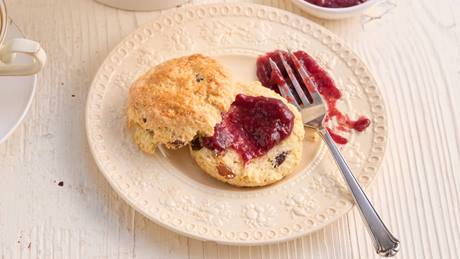 Auf einer weißen Tortenplatte liegen mehrere Scones mit Rosinen. Ein Scone liegt aufgeschnitten davor auf einem weißen Kuchen-Teller. Die eine Hälfte des Scone ist mit einer roten Konfitüre bestrichen, daneben liegt eine Kuchengabel. Links neben dem Kuchenteller seht eine weiße verschnörkelte Tasse mit Tee.