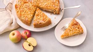 Auf einem großen weißen Porzellan-Teller ist ein angeschnittener Apfelkuchen mit Tortenheber. Ein Stück des Kuchens liegt mit einer Gabel auf einem separaten kleinen Kuchen-Teller. Daneben liegen drei Äpfel, einer davon halbiert.