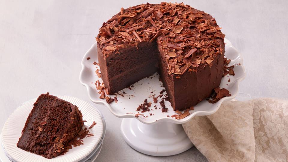 Ein brauner Schokoladen-Kuchen steht auf einem weißen Tortenständer. Ein Stück des Kuchens fehlt. Zwei Teller stehen ebenfalls auf dem Tisch, jeder mit einem Stück des Schoko-Kuchens. Auf jedem Teller liegt eine Gabel. Der Kuchen ist mit Schokoladenraspeln bestreut.