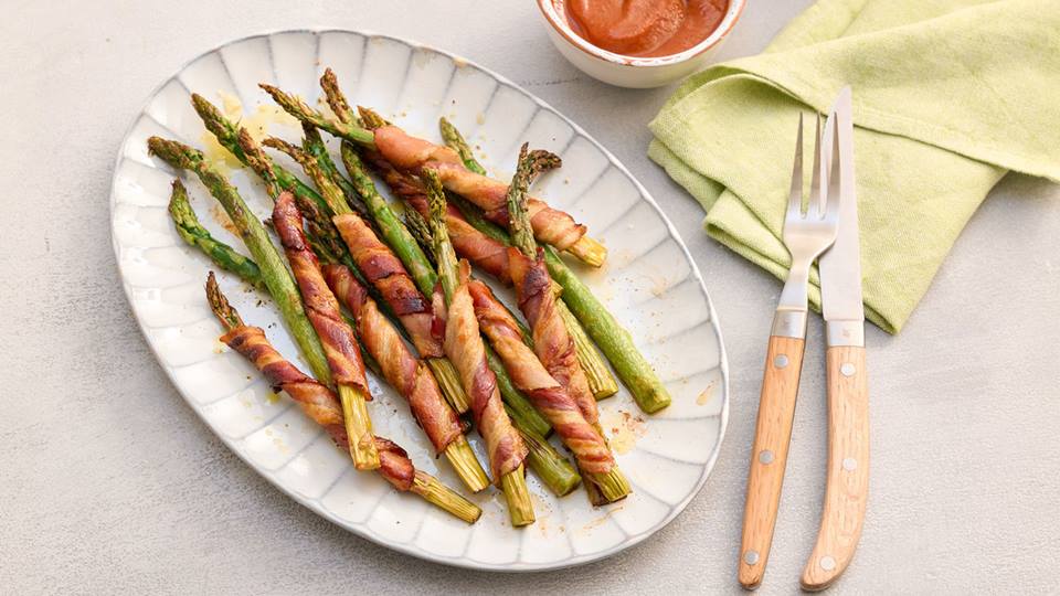 Grüner Spargel, teils mit Speck umwickelt, auf weißem Teller mit Messer und Gabel neben grünem Stoffserviette und Schälchen mit roter Sauce.