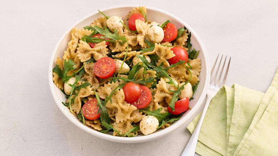 Schale mit Nudelsalat aus Farfalle, Kirschtomaten, Mozzarellakugeln und Rucola neben einer Gabel und einem hellgrünen Stoffserviette.