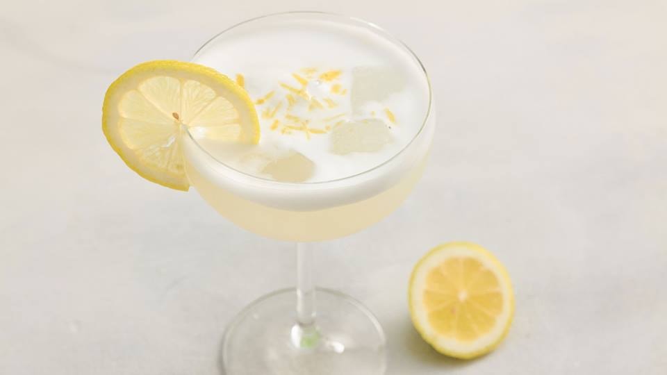 Cocktailglas mit einem hellen Getränk, Eiswürfeln, einer Zitronenscheibe am Glasrand und einer halben Zitrone danebe.