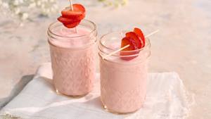 Zwei Gläser mit rosa Erdbeer-Lassi, jeweils mit einer aufgespießten Erdbeerscheibe als Garnitur, auf weißem Tuch.