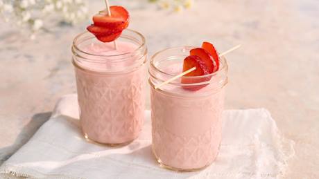 Zwei Gläser mit rosa Erdbeer-Lassi, jeweils mit einer aufgespießten Erdbeerscheibe als Garnitur, auf weißem Tuch.