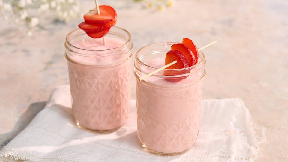 Zwei Gläser mit rosa Erdbeer-Lassi, jeweils mit einer aufgespießten Erdbeerscheibe als Garnitur, auf weißem Tuch.