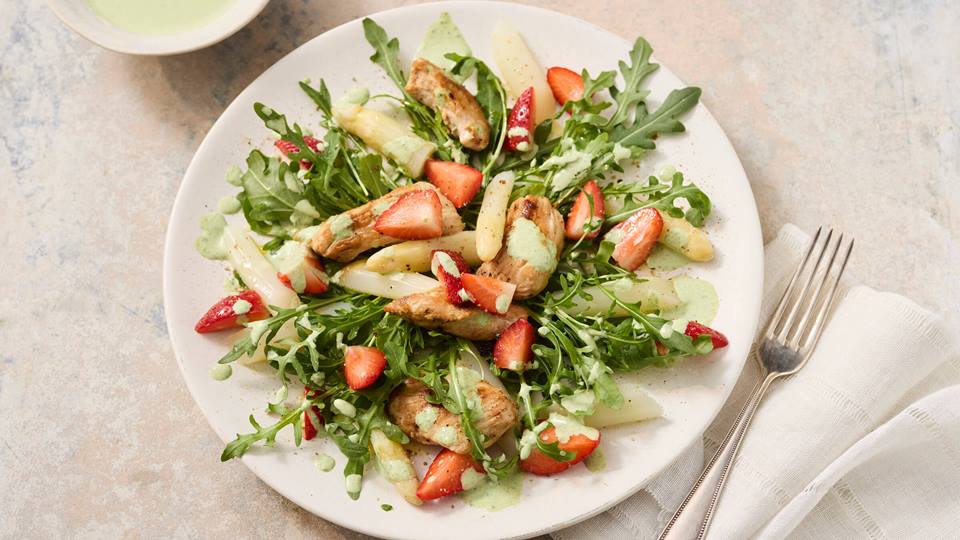 Teller mit Salat aus weißem Spargel, Erdbeeren, Rucola und Hähnchenstreifen, daneben eine Gabel auf weißem Stoffserviette.