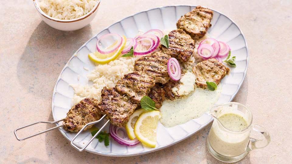 Gegrillte Fleischspieße auf weißem Teller mit Reis, roten Zwiebelringen, Zitronenscheiben, Minzjoghurt und Minzblättern, daneben kleine Schale mit Reis und Glas mit Minzjoghurtsoße.