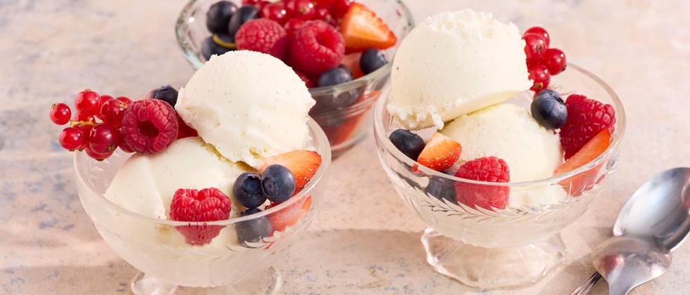 Zwei Gläser mit je einer Kugel Vanilleeis und Beeren sowie eine Schale mit gemischten Beeren auf hellem Untergrund