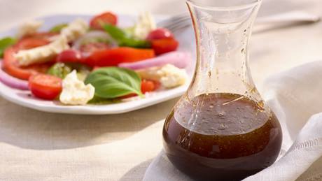 Glaskaraffe mit Balsamico Dressing vor einem Teller mit Tomaten, Mozzarella und Basilikum auf hellem Tischtuch.