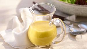 Glaskrug mit klassischer Vinaigrette auf weißem Tuch vor Salatschüssel und Besteck.