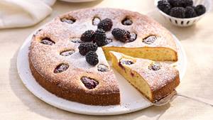 Runder Kuchen mit Puderzucker bestäubt, mit Brombeeren belegt, ein Stück wird mit einem Kuchenheber herausgehoben