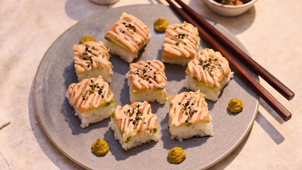 Neun Sushi-Quadrate mit rosa Sauce und Sesam auf grauem Teller, daneben Essstäbchen und kleine grüne Wasabihäufchen.