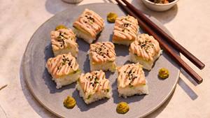 Neun Sushi-Quadrate mit rosa Sauce und Sesam auf grauem Teller, daneben Essstäbchen und kleine grüne Wasabihäufchen.