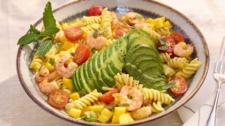 Schale mit Fusilli-Nudeln, Garnelen, halbierten Kirschtomaten, gewürfelter Mango, Avocadoscheiben und Minzblatt auf hellem Tuch