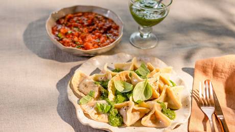 Teller mit Ravioli, garniert mit grünem Pesto und Basilikumblättern, daneben Schale mit Tomatensauce und ein Glas grünes Pesto mit Teelöffel auf hellem Tuch