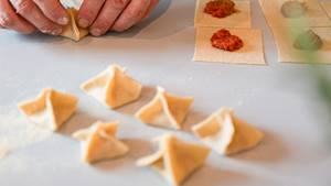 Hände falten Teigquadrate mit roter und grüner Füllung zu kleinen Teigtaschen auf hellem Untergrund