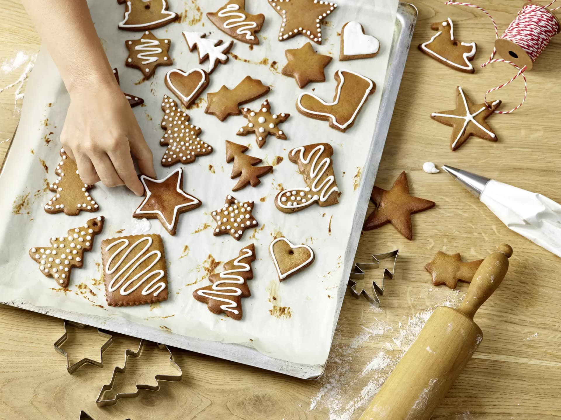 Lebkuchen in verschiedenen Formen liegen auf einem mit Backpapier ausgelegten Blech. Die Lebkuchen sind mit weißem Zuckerguss verziert, zum Beispiel als Sterne, Tannenbäume, Herzen und Stiefel. Eine Hand greift nach einem der Lebkuchen. Auf dem Holztisch daneben liegen Ausstechformen, ein Nudelholz, etwas Mehl und eine Spritztüte mit weißem Zuckerguss. Im Hintergrund ist eine Rolle mit rot-weißem Bäckergarn zu sehen.