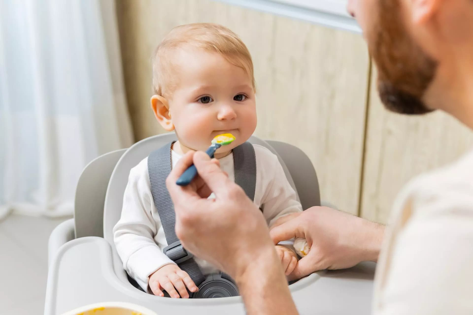 Ein Baby sitzt sehr ruhig in einem Hochsitz und bekommt orange farbenen Brei mit einem blauen Löffel von einem Mann mit Bart gefüttert.
