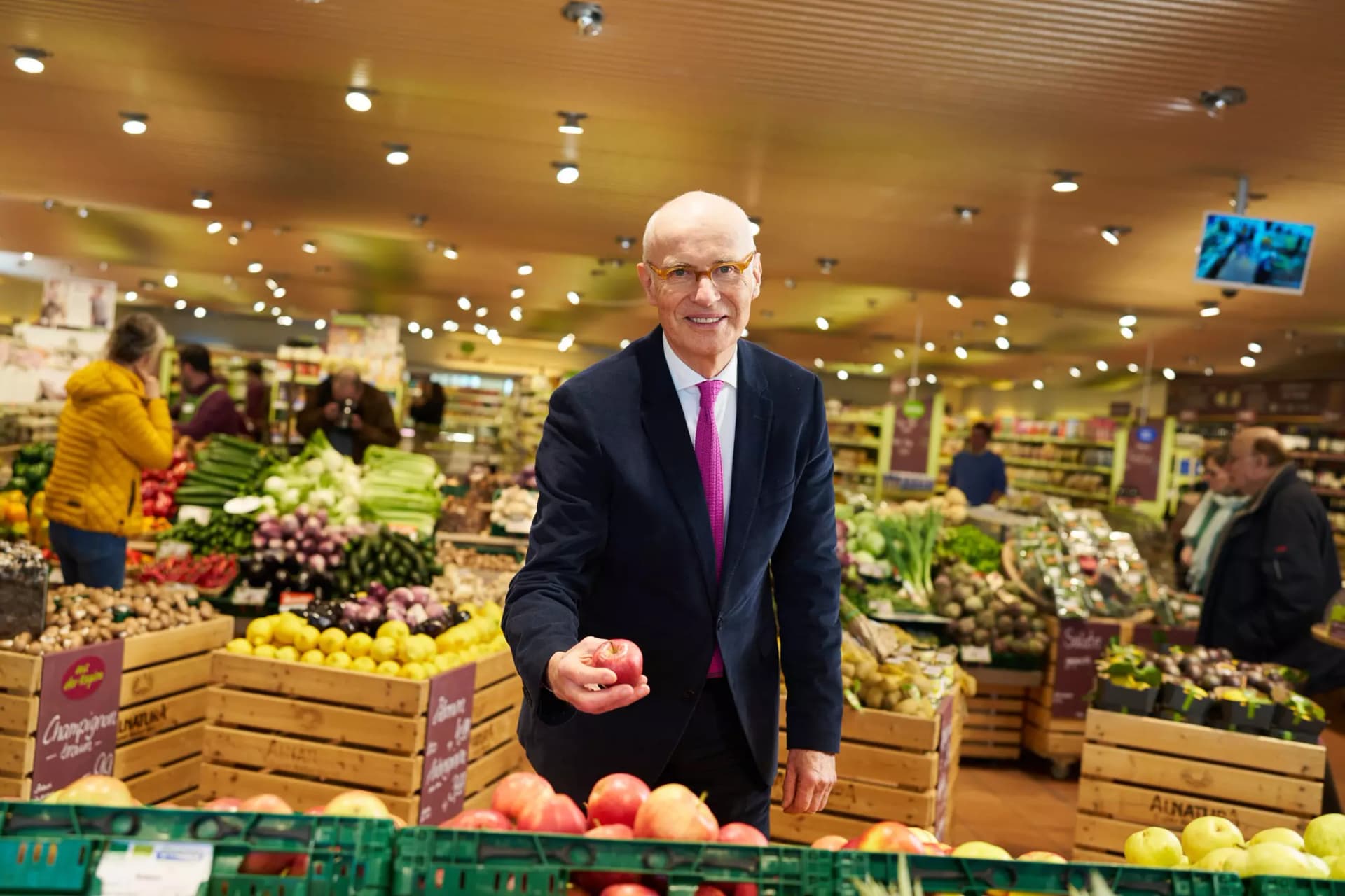 Alnatura Unternehmensgründer Götz E. Rehn steht in einem Alnatura Super Natur Markt an der Obsttheke und hält einen Apfel.