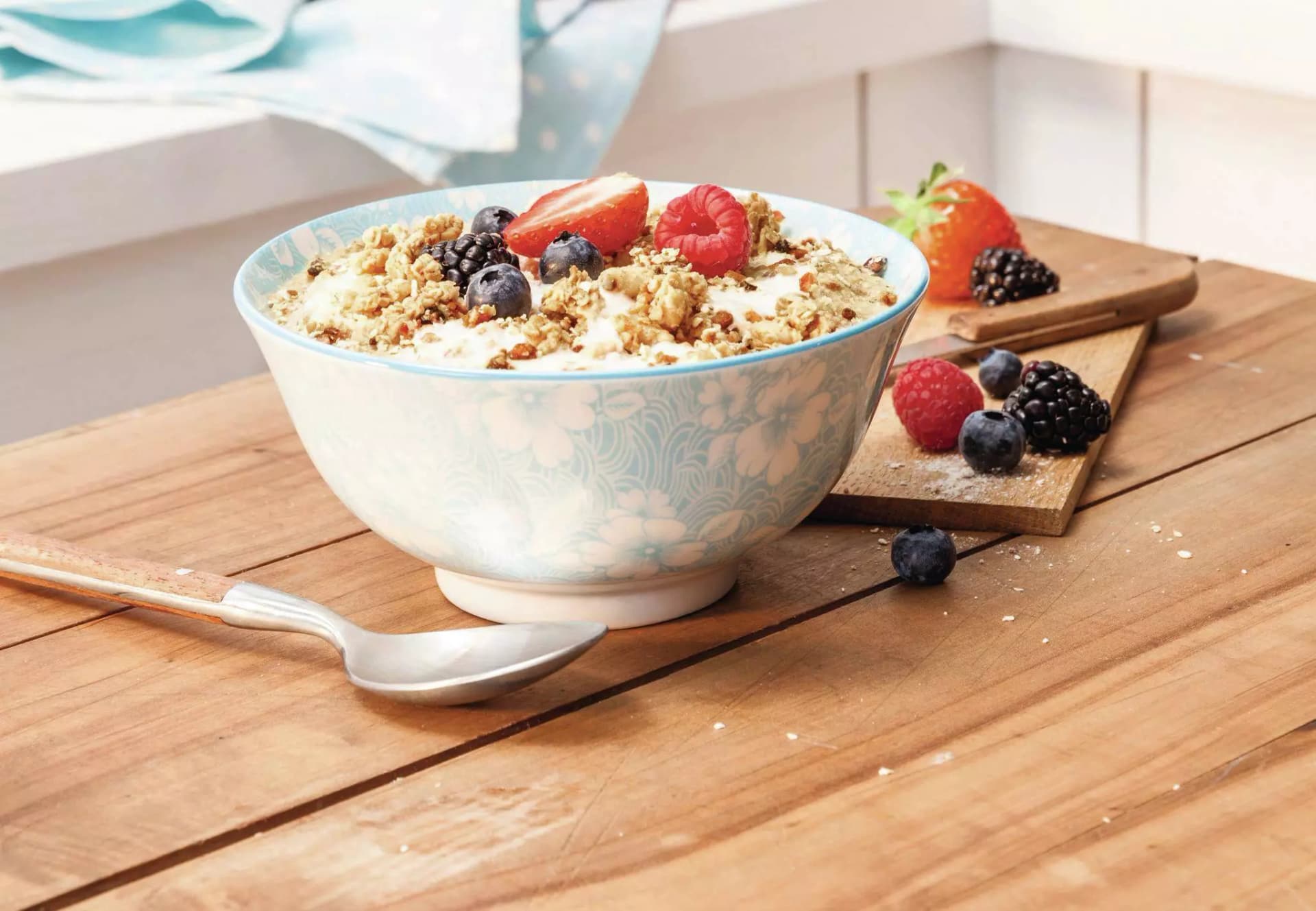 Schale mit Müsli, Joghurt und Beeren auf Holztisch neben Löffel und Schneidebrett mit weiteren Beeren.