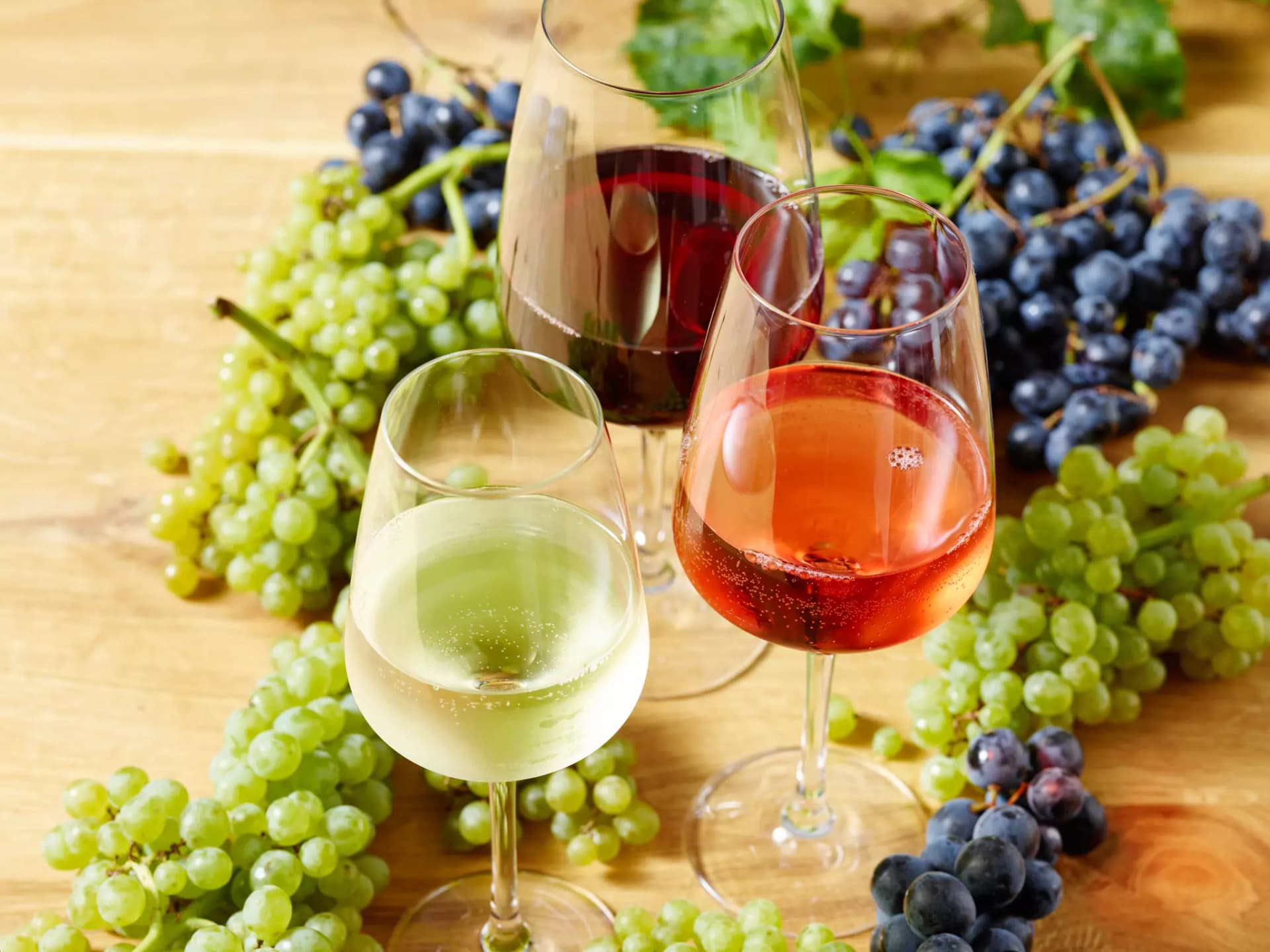 Drei Weingläser mit Weißwein, Roséwein und Rotwein vor Trauben auf Holztisch.
