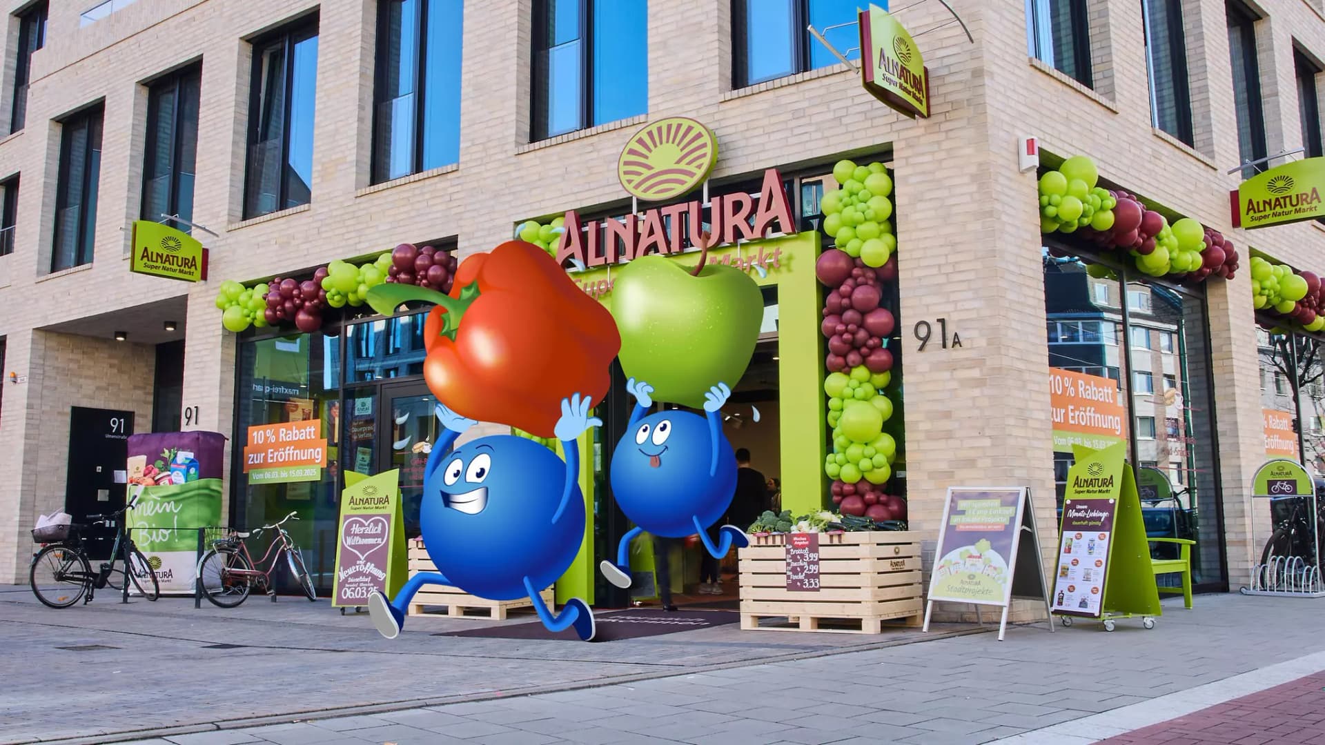 Eingang eines Alnatura-Geschäfts in einem modernen Gebäude mit bunten Luftballons und zwei großen blauen Comicfiguren, die einen roten Apfel und eine grüne Birne halten und aus dem Markt laufen.