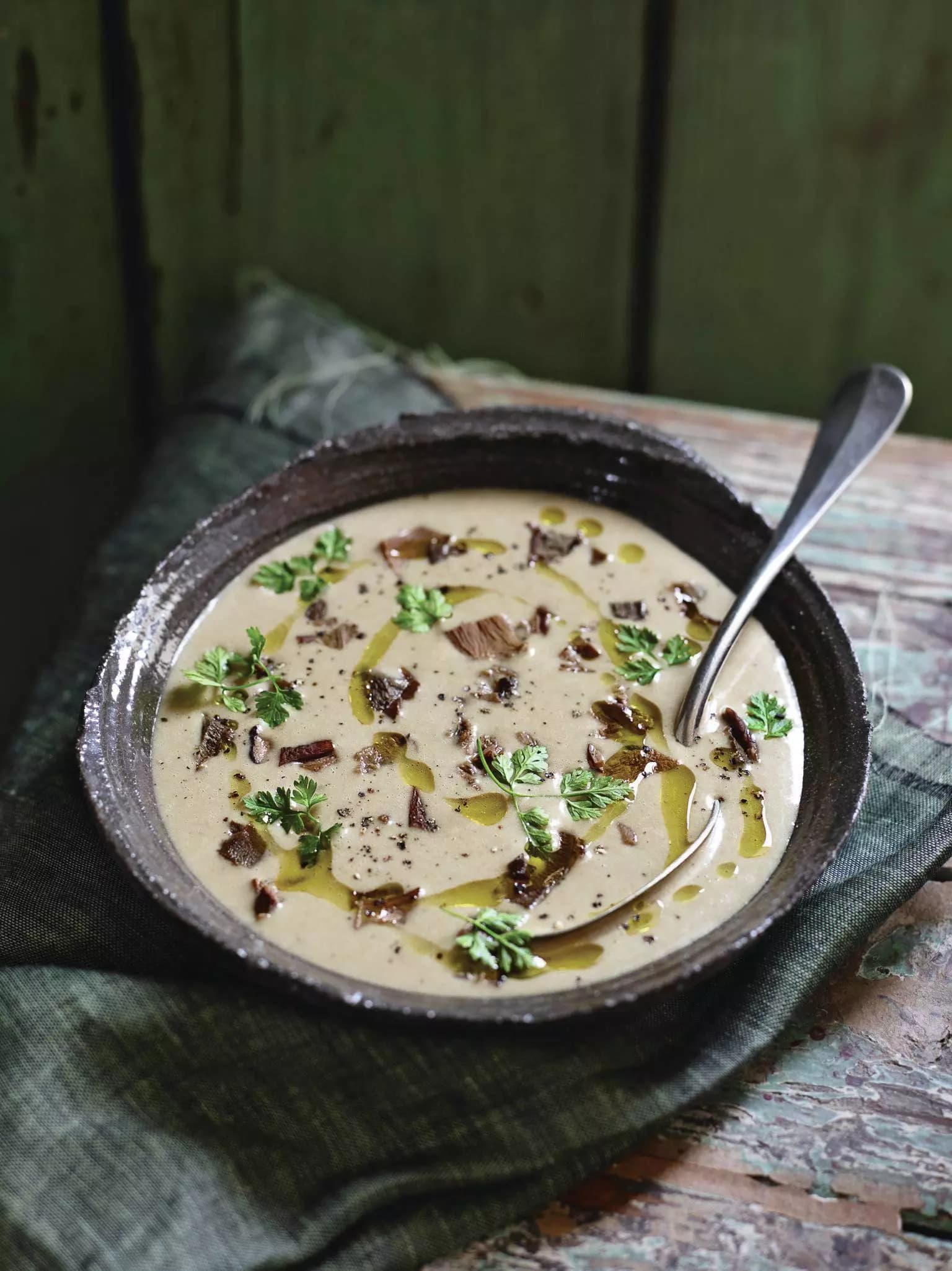 Schale mit cremiger Suppe, garniert mit Kräutern, Olivenöl und kleinen Pilzstücken, auf einem dunklen Tuch und einem rustikalen Holztisch