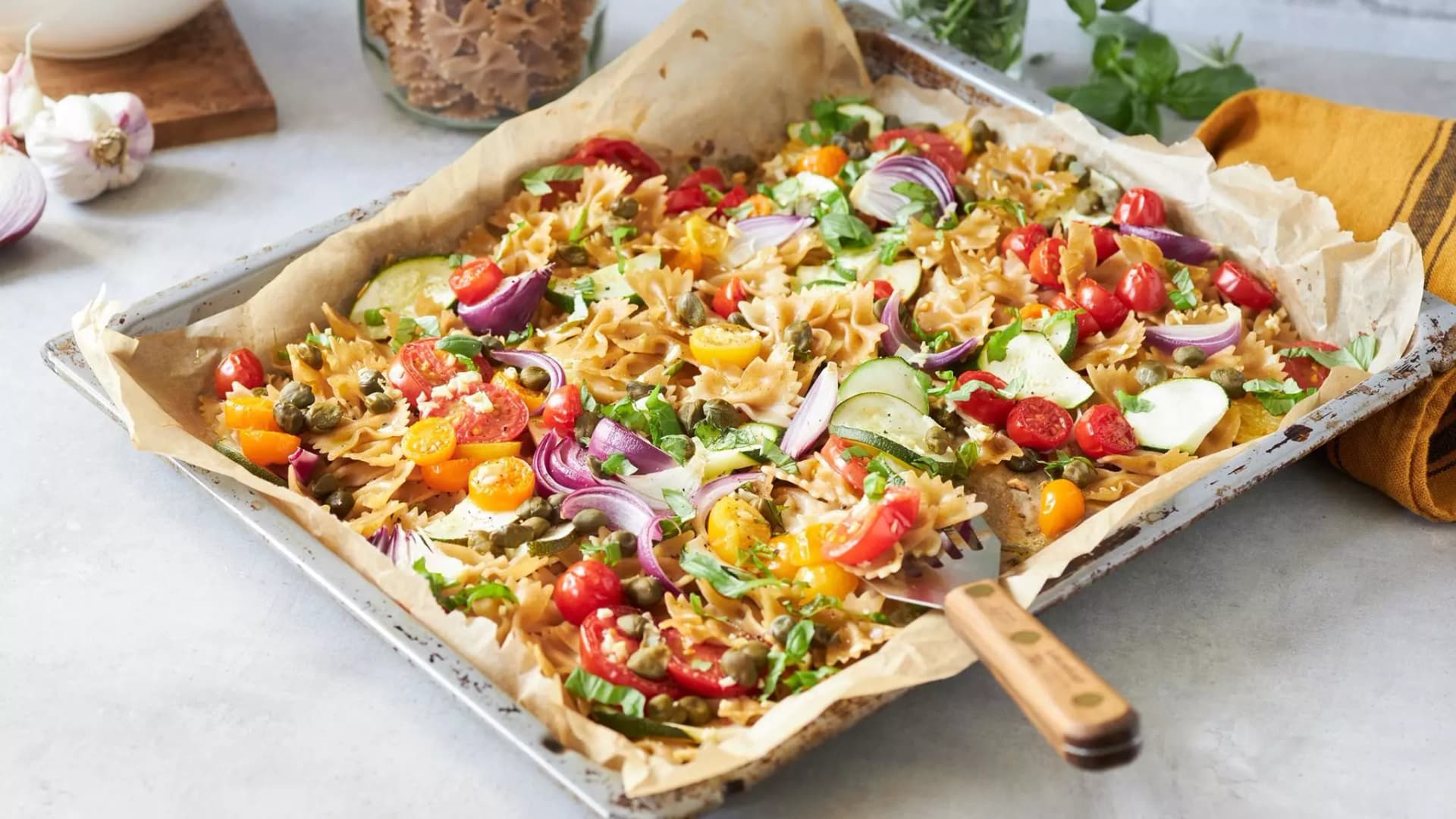 Backblech mit bunter Farfalle-Pasta, Kirschtomaten, Zucchinischeiben, roten Zwiebelringen und Kapern auf Backpapier