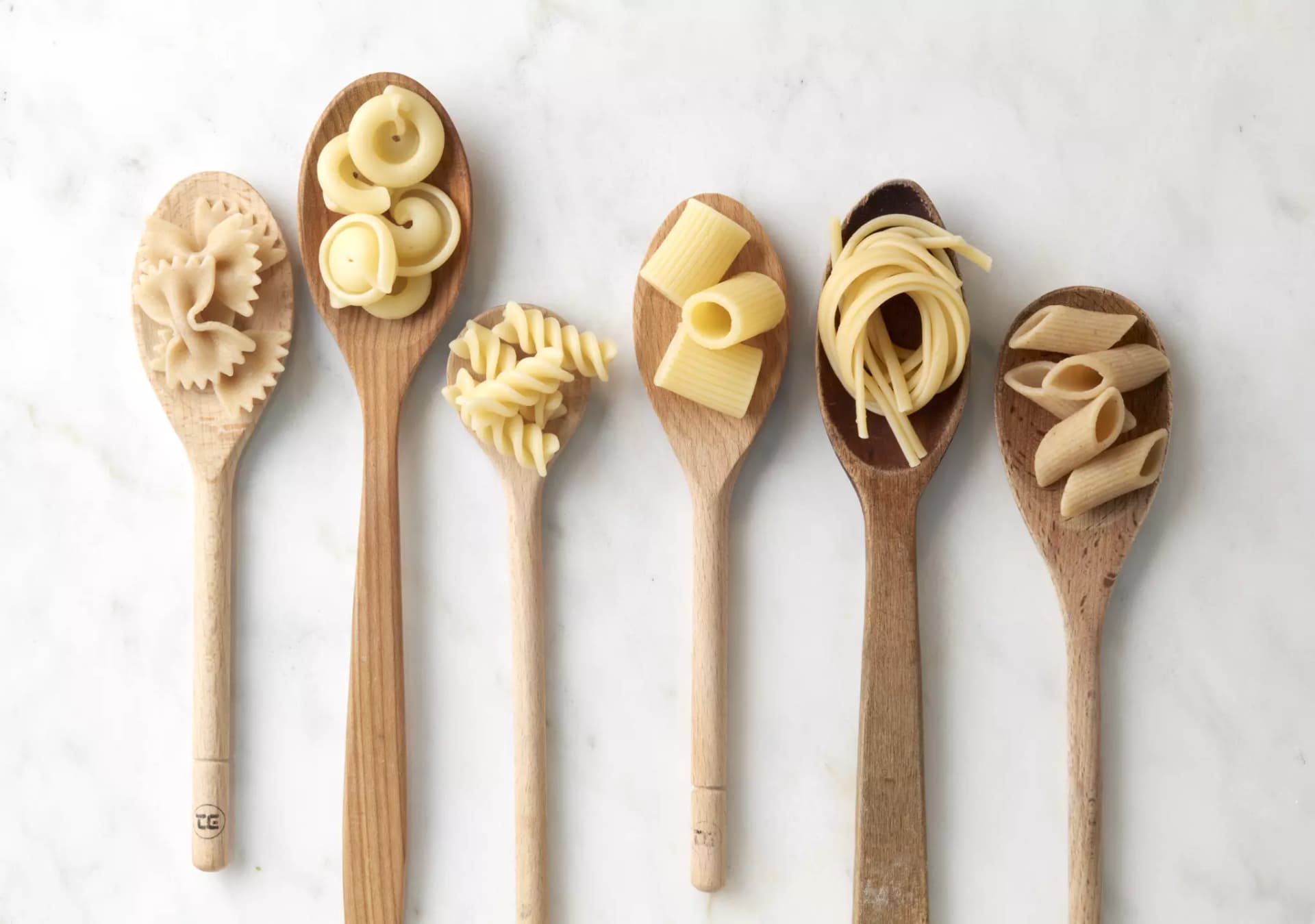 Sechs liegende Holzkochlöffel, in jeder Mulde sind andere Pastasorten.