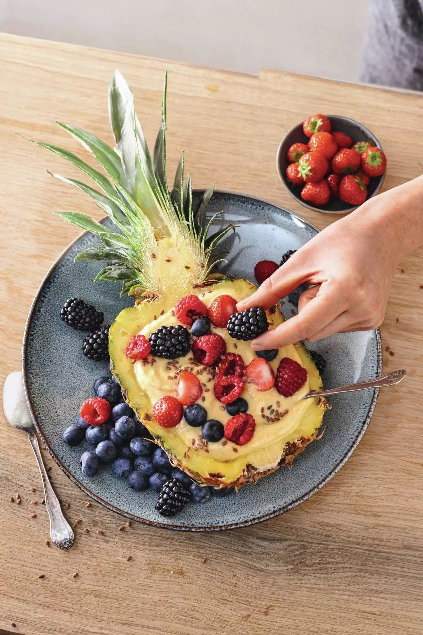 Halbierte Ananas mit Joghurt und verschiedenen Beeren auf einem Teller, daneben eine Schale mit Erdbeeren und eine Hand, die Beeren auf die Ananas legt.