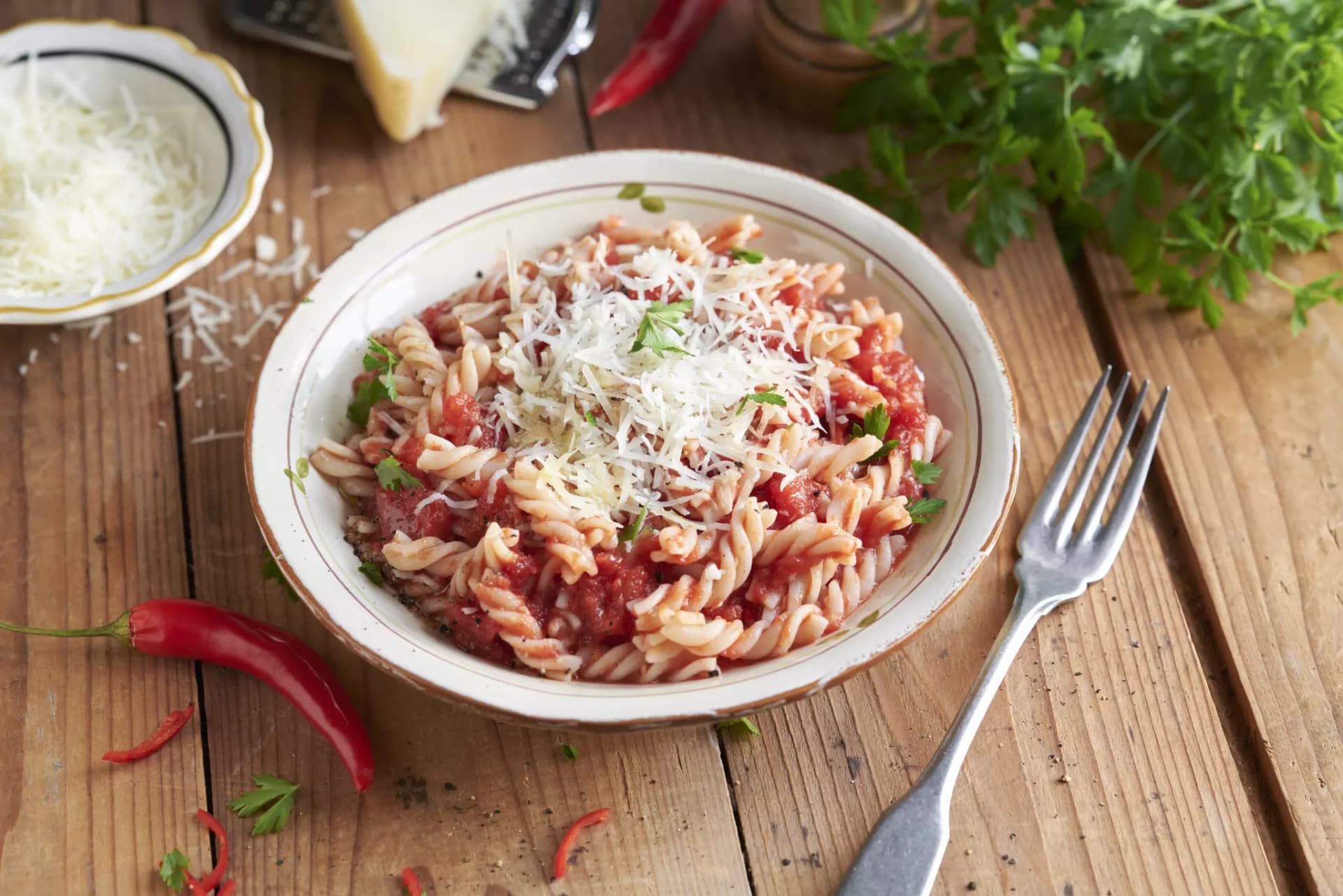 Teller mit Fusilli-Pasta in Tomatensauce, bestreut mit geriebenem Käse und Petersilie, daneben rote Chilischoten und eine Gabel auf Holzuntergrund.