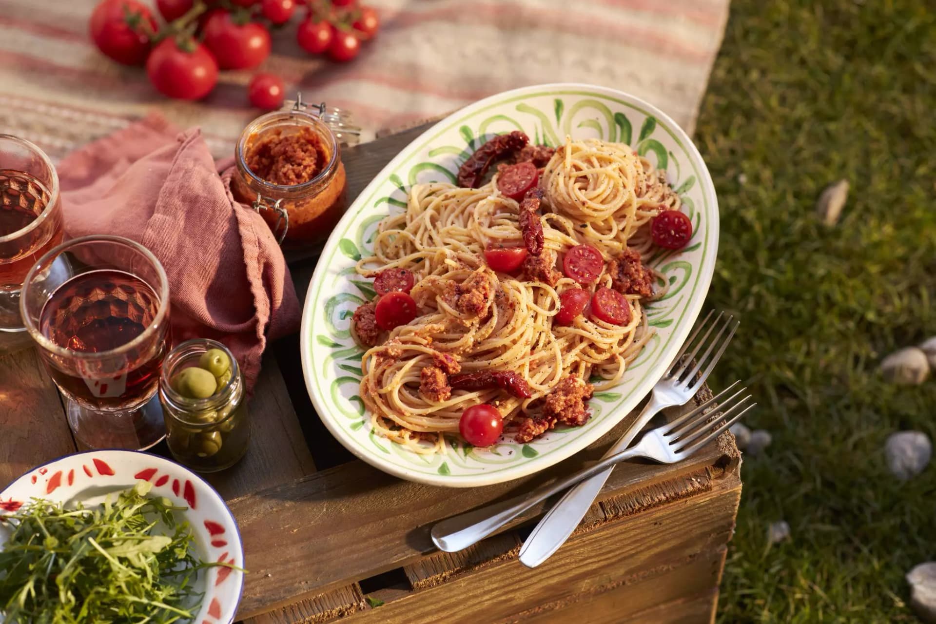 Teller mit Spaghetti, Tomatensauce, Kirschtomaten und getrockneten Tomaten auf einem Holztisch mit Besteck, Glas mit Getränk, Glas mit Oliven und einem Glas mit Tomatensauce daneben.