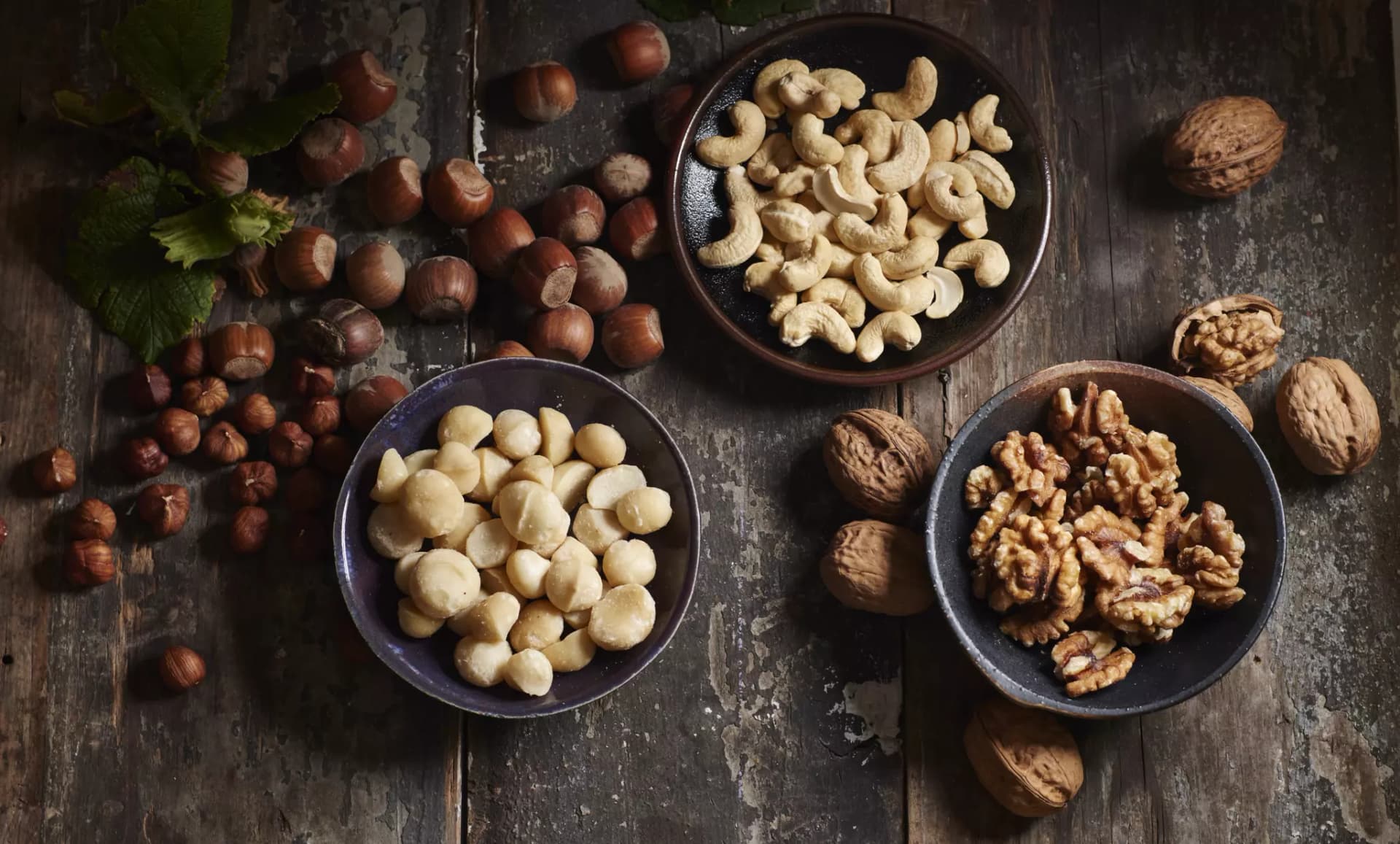 Drei Schalen mit Cashewkernen, Haselnüssen und Walnusskernen auf dunklem Holztisch mit verstreuten ganzen Nüssen und Haselnusszweigen.