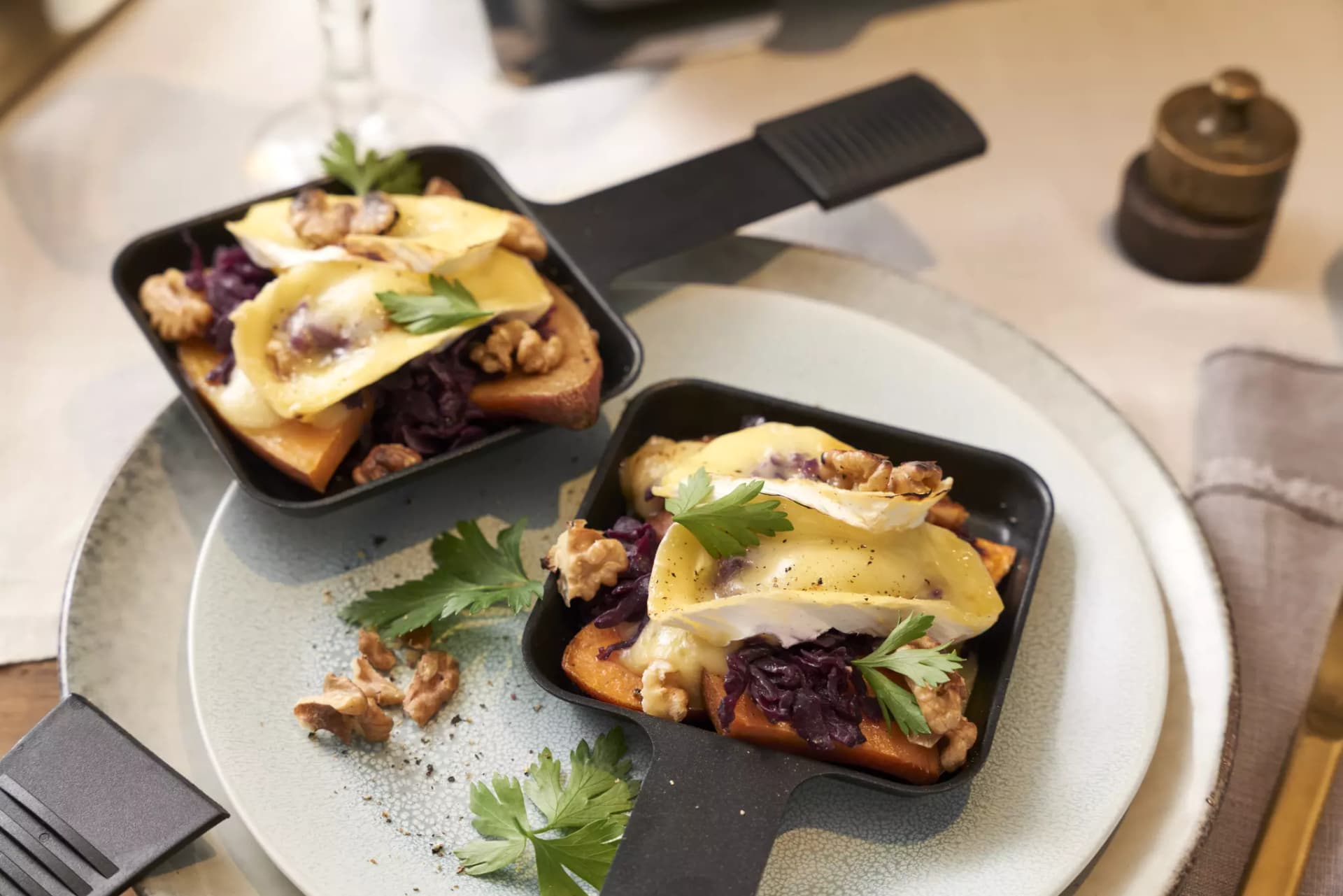 Zwei kleine schwarze Pfannen mit Toast, Walnüssen, Käse, roten Zwiebeln und Petersilienblättern auf einem grauen Teller.